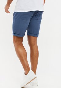 Mørkeblå denim shorts med rullekanter, der har baglommer og en skræddersyet pasform, parret med hvide sneakers på en ensfarvet baggrund.