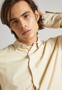 Camicia beige con bottoni, realizzata in un tessuto morbido e con un motivo testurizzato, dotata di colletto classico. Presenta bottoni bianchi e una vestibilità rilassata.