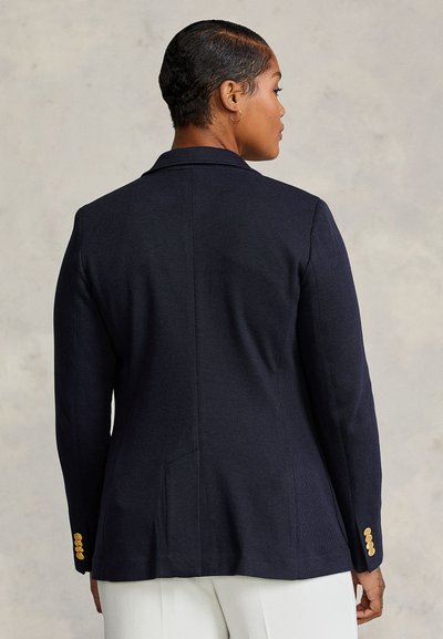 Femme aux cheveux courts portant un blazer bleu marine avec des boutons dorés sur les manches et un pantalon blanc, face à un fond gris clair.