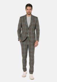 Mann in grauem Karosakkos mit hellgrauem Hemd und weißen Sneakers, der vor einem schlichten weißen Hintergrund steht.