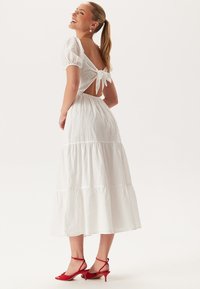 Witte midi-jurk met puffmouwen, strikdetail op de rug, gelaagde rok, gestructureerde stof en rode sandaaltjes met bandjes.