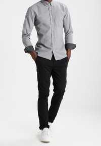 Camisa abotonada gris con cuello, puños negros contrastantes y botones marrón claro; combinada con pantalones negros y zapatillas blancas.
