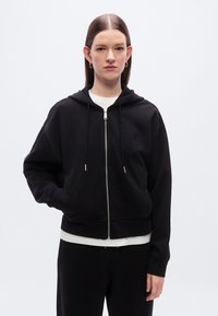 Μαύρο φούτερ με φερμουάρ και κουκούλα με κορδόνι, με ριμπ μανσέτες και τελείωμα. Απαλό υλικό, με λογότυπο στο στήθος, συνδυασμένο με λευκή μπλούζα από κάτω.