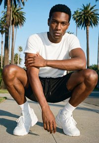 Jonge man in een wit T-shirt, zwarte korte broek en witte sneakers hurkend op het trottoir met palmbomen en een blauwe lucht op de achtergrond.