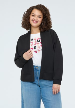 Smilende kvinde med krøllet hår iført sort åben jakke, hvid t-shirt med grafik og blå jeans foran en ensfarvet lys baggrund.