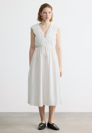 Νεαρό άτομο όρθιο, φορώντας ένα λευκό αμάνικο φόρεμα μήκους μέχρι τη μέση της γάμπας με δέσιμο μπροστά και μαύρα παπούτσια Mary Jane, σε γκρι πάτωμα.