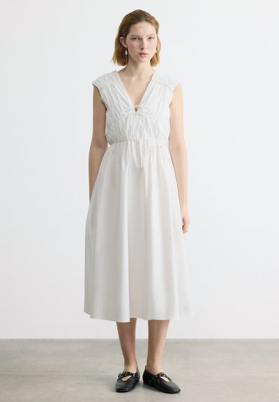 Jeune personne debout en posture droite portant une robe midi blanche sans manches avec un nœud devant et des chaussures Mary Jane noires sur un sol gris.