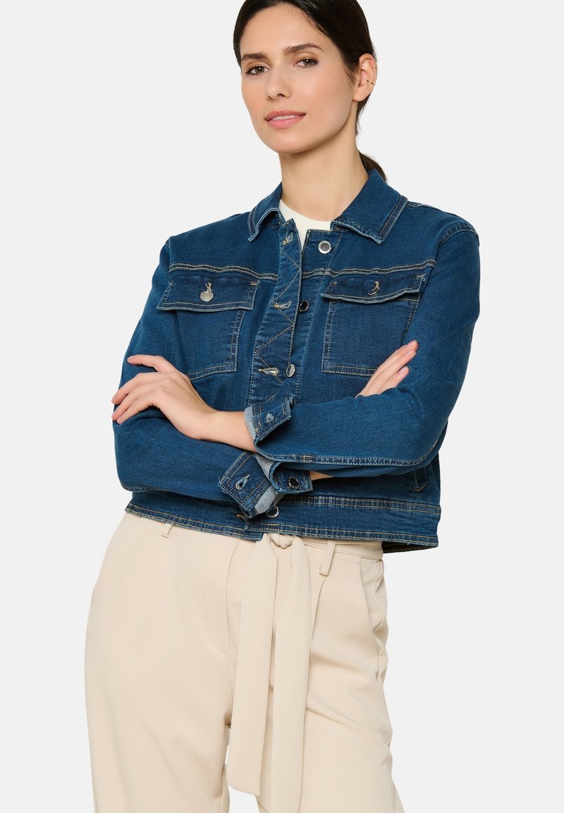 Tamsiai mėlynas džinsinis švarkas su dviem krūtinės kišenėmis, sidabriniais mygtukais ir trumpu dizainu. Dėvimas virš šviesiai smėlio spalvos aukšto liemens kelnių.