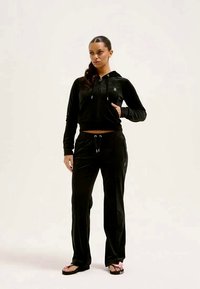 Melnā zīda treniņtērps ar rāvējslēdzēju un šņorējamām biksēm. Tam ir neliels logotips uz krūtīm, gluda tekstūra un brīva piegriezuma dizains.