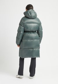 Lange, gewatteerde pufferjas met kap in een matte, donkerblauwe kleur. Kenmerken zijn een getailleerde taille en geborduurde secties. Draag het over zwarte broeken en lichte schoenen.