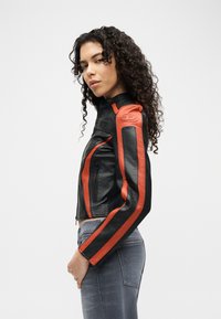 Νεαρή γυναίκα με σγουρά μαλλιά που φοράει μαύρο δερμάτινο μπουφάν με κόκκινες ρίγες και γκρι τζιν, στέκεται πλάγια μπροστά σε απλό φόντο.