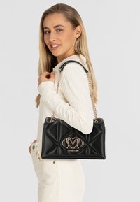 Czarna pikowana torebka wykonana z syntetycznej skóry, z złotym łańcuszkiem i ozdobnym logo z przodu. Strukturalny design i gładka faktura.