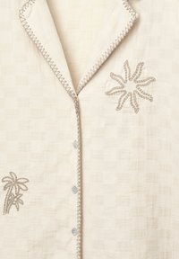 Chemise beige clair en tissu texturé, avec un col, des boutons bleus, et des motifs brodés de palmiers et de fleurs en fil taupe.