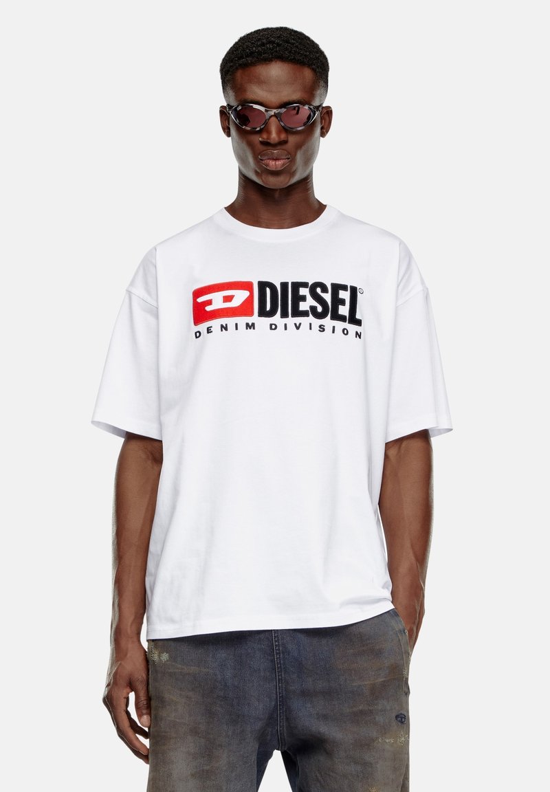 Biała bawełniana koszulka z dużym czerwonym i czarnym logo "DIESEL" oraz napisem "DENIM DIVISION". Model nosi ciemne okulary przeciwsłoneczne w połączeniu z wyblakłymi dżinsami.