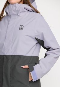 Lichtpaarse en donkergrijze waterdichte jas met een hoge kraag, voorzien van een logo, zakken en klittenbandsluitingen bij de mouwen.