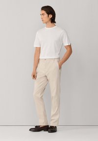 Uomo che indossa una t-shirt bianca a maniche corte, pantaloni beige e mocassini marrone scuro con nappine, in piedi con una mano in tasca, guarda verso sinistra.