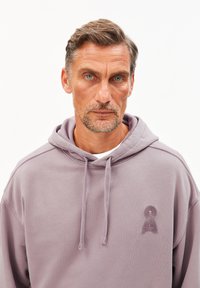 Uomo di mezza età con una leggera barba e occhi azzurri, che indossa una felpa con cappuccio viola chiaro con un piccolo logo ricamato sul petto, sfondo neutro.