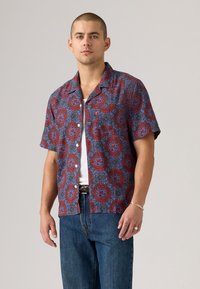Young man wearing a short-sleeve red and blue patterned button-up shirt over a white tank top and blue jeans, standing against a plain background.