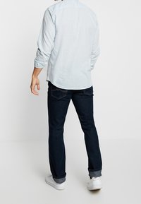 Světle modrá košile s dlouhým rukávem a s ohrnutými rukávy, v kombinaci s tmavě modrými džíny a bílými teniskami, viděná zezadu.