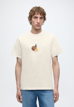 Jeune homme aux cheveux clairs et bouclés portant un t-shirt crème avec un motif de basket-ball, une bouteille de vin et deux citrouilles imprimés sur la poitrine.