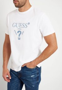 T-shirt en coton blanc avec un imprimé graphique bleu portant les mentions "GUESS" et "WASHED JEANS". Le t-shirt a un col rond et des manches courtes.