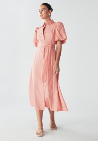 Vestido rosa de botones con mangas abullonadas y cintura ceñida, con una silueta fluida y dobladillo hasta la rodilla. Textura de tela suave.