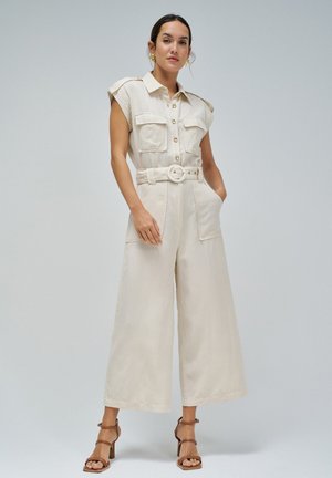 Femme portant une combinaison beige sans manches avec des boutons sur le devant, taille ceinturée, jambes larges, et sandales à talons marron, debout sur un fond gris.