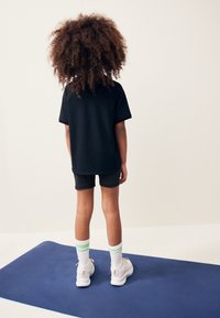 Czarna koszulka z krótkim rękawem, ciemne szorty, białe sneakersy z jasnymi akcentami, skarpetki w zielone paski. Stoi na niebieskiej macie, widok z tyłu.