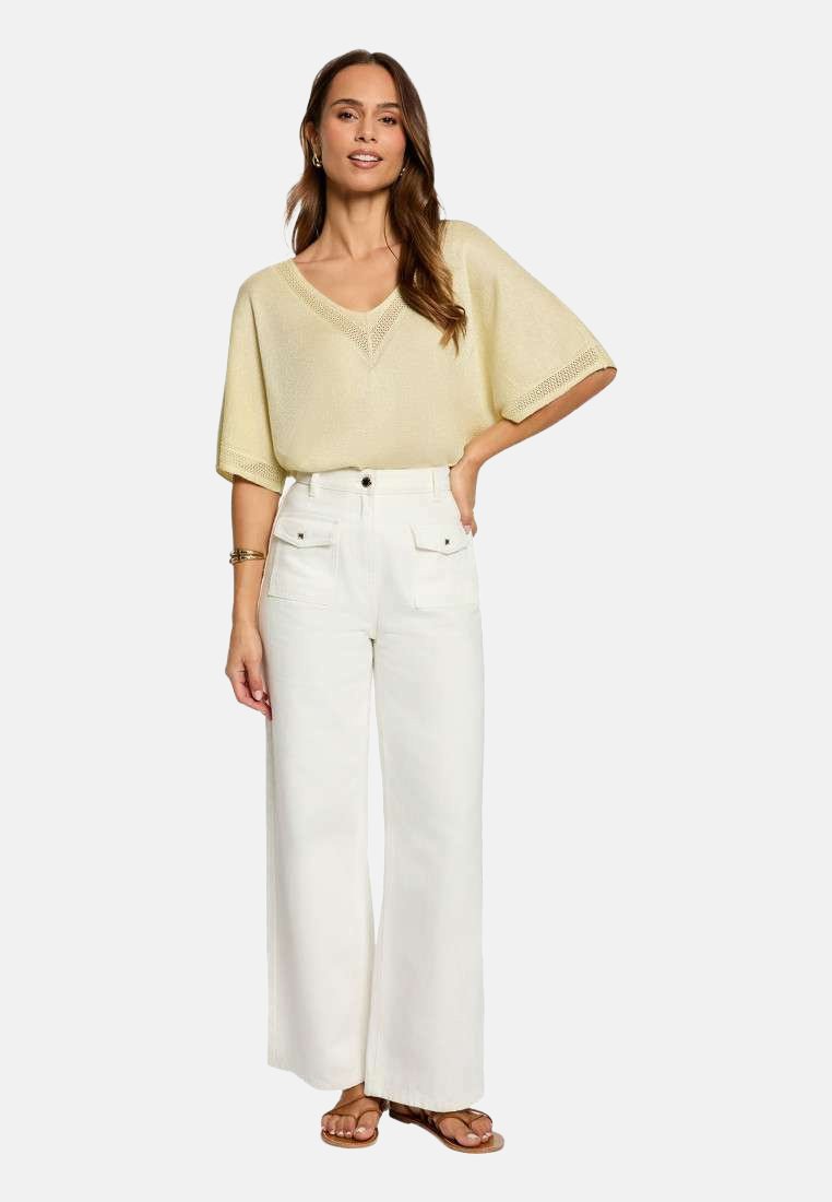 Femme portant un haut en tricot à manches courtes jaune clair, un pantalon blanc taille haute à jambes larges avec poches devant, et des sandales marron, debout avec une main sur la hanche.