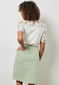Falda de mezclilla verde menta con dos bolsillos traseros, combinada con una camiseta blanca con recortes florales y mangas cortas.