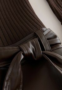 Top maro din tricot cu dungi, cu o curea din piele ecologică la talie, evidențiind un detaliu de fundă legată și o textură netedă, subliniind materialele contrastante.