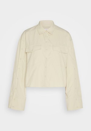 Cropped, cream-colored shirt in lightweight fabric. Features a pointed collar, two chest pockets, and button details along sleeve edges.