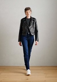 Črna usnjena motoristična jakna s srebrnimi dodatki, v kombinaciji s temno modrimi kavbojkami in belimi športnimi copati. Model stoji na leseni talni površini ob enobarvni steni.