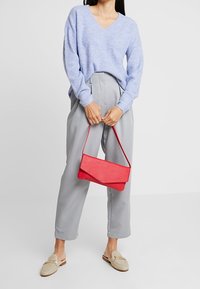 Osoba ve světle modrém svetru a šedých kalhotách, která drží červenou kabelku a má na sobě béžové boty typu slip-on.