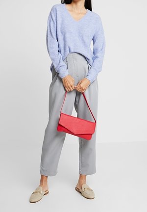 Persoon in een lichtblauwe trui en grijze broek, met een rode handtas in de hand en beige instappers zichtbaar.