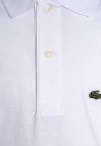 Biała koszulka polo wykonana z bawełny, z kołnierzykiem, dwoma widocznymi guzikami oraz zielonym wyszywanym logo po lewej stronie.