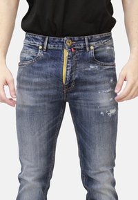 Jeans in denim blu con un finish rovinato, dotati di cerniera gialla, bottone in ottone e cinque tasche. Cuciture blu accentuano il design.