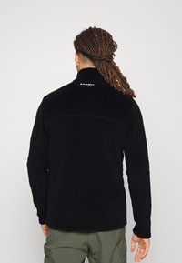 Black fleece jacket with a high collar, subtle stitching details, and a logo on the upper back. Soft texture and relaxed fit.