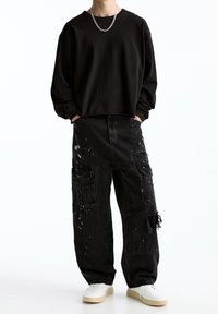 Camisa negra de manga larga, corte holgado. Pantalones cargo negros con múltiples bolsillos, detalles de salpicaduras de pintura y bordes desgastados. Zapatillas blancas.