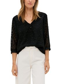 Zwarte blouse met een gestructureerd luipaardpatroon, driekwart doorzichtige mouwen, V-hals met knoop, gecombineerd met lichtgekleurde broeken.