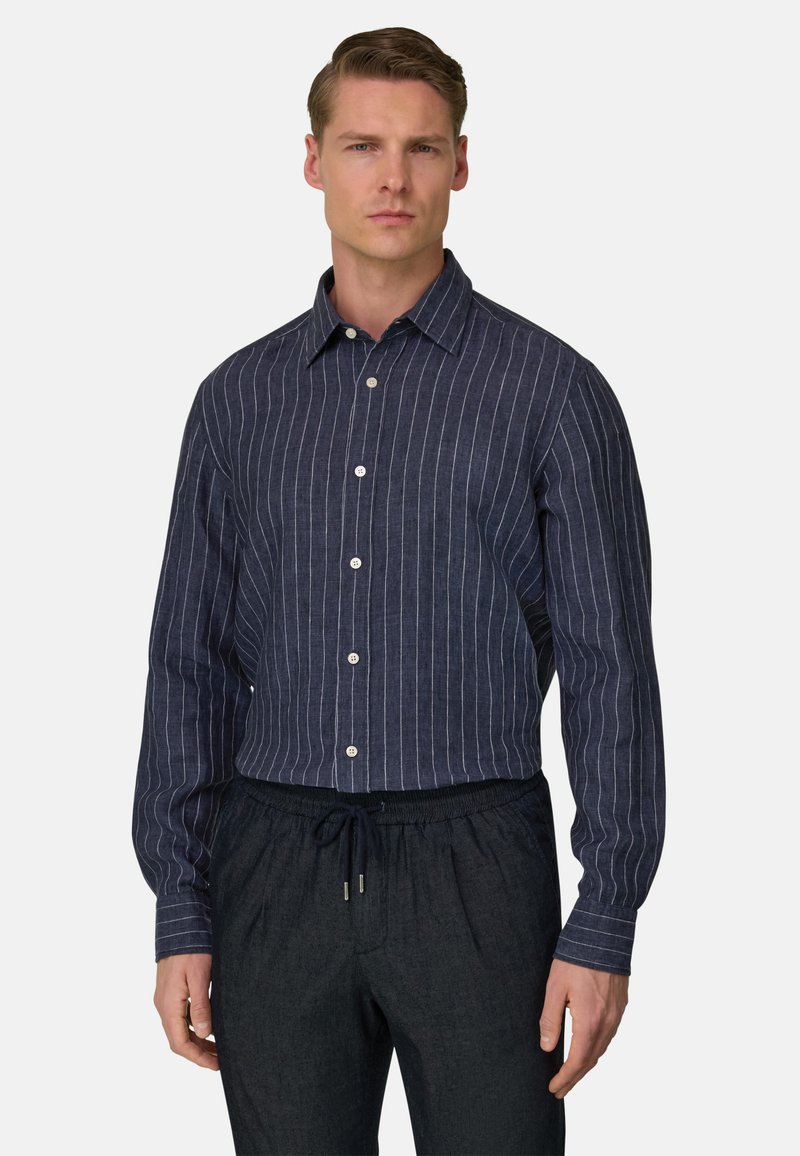 Homme portant une chemise à manches longues bleu marine à rayures verticales blanches et un pantalon à cordon de serrage foncé, debout devant un fond uni.