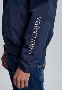 Chaqueta bomber de color azul marino con puños elásticos, que cuenta con un bolsillo con cremallera y un logo en color beige claro en la manga. Material suave y ligero.