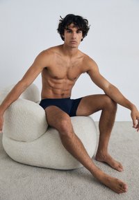 Mørk marineblå badebukser på en sittende mannlig figur med muskuløs fysikk, posert på en strukturert, off-white, rund stol. Lys bakgrunn.