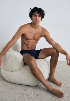 Tmavomodré plavky na sedejúcej mužskej postave s vyrysovanou postavou, zasadené na textúrovanej, krémovej, zaoblenej stoličke. Svetlé pozadie.