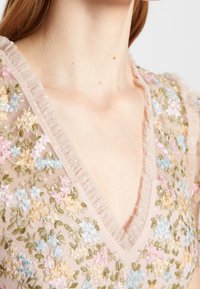 Blusa ricamata con scollatura a V, bordi delicati a volant e motivi floreali in rosa, blu e verde su un tessuto nudo e trasparente.