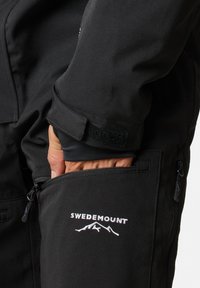 Zwarte softshell jack met een ritszak aan de zijkant, klittenband manchetten en een wit logo met bergafbeeldingen. Glad textuur.