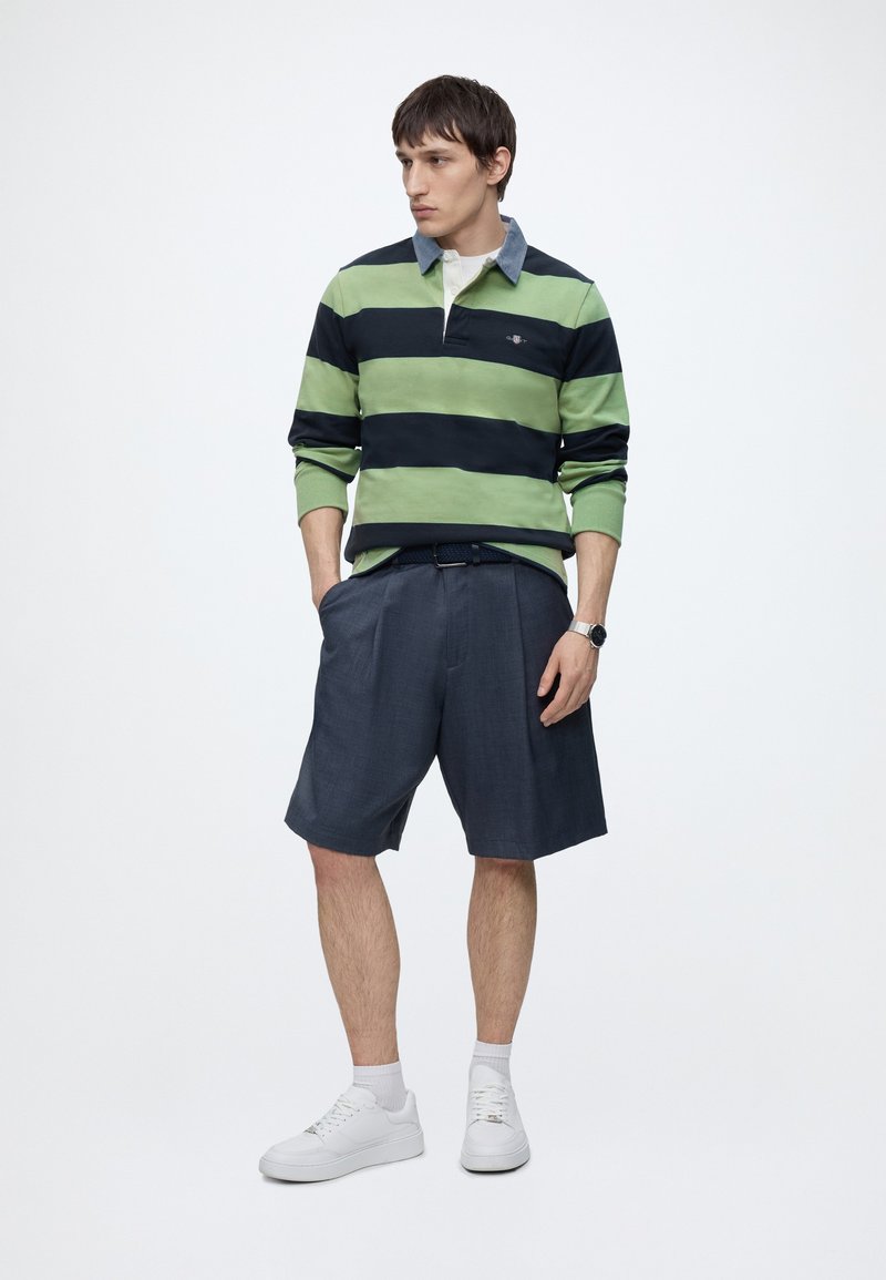 Un homme debout porte un polo de rugby à rayures vertes et bleu marine, un short sombre et ample, des chaussettes blanches et des baskets, les mains dans les poches, regardant vers la gauche.