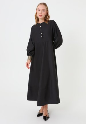 Kvinde iført en lang sort kjole med knapdetaljer på brystet, puffede lange ærmer, sorte spidse sko og et guld manchetarmbånd.