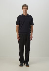 Polo shirt navy avec un col, associé à un pantalon de sport noir avec des rayures blanches sur les côtés. Les chaussures sont noires avec une finition texturée.