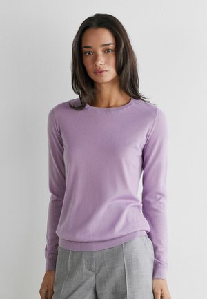 Femme aux longs cheveux foncés portant un pull lavande à manches longues et un pantalon gris taille haute, debout devant un mur blanc uni.